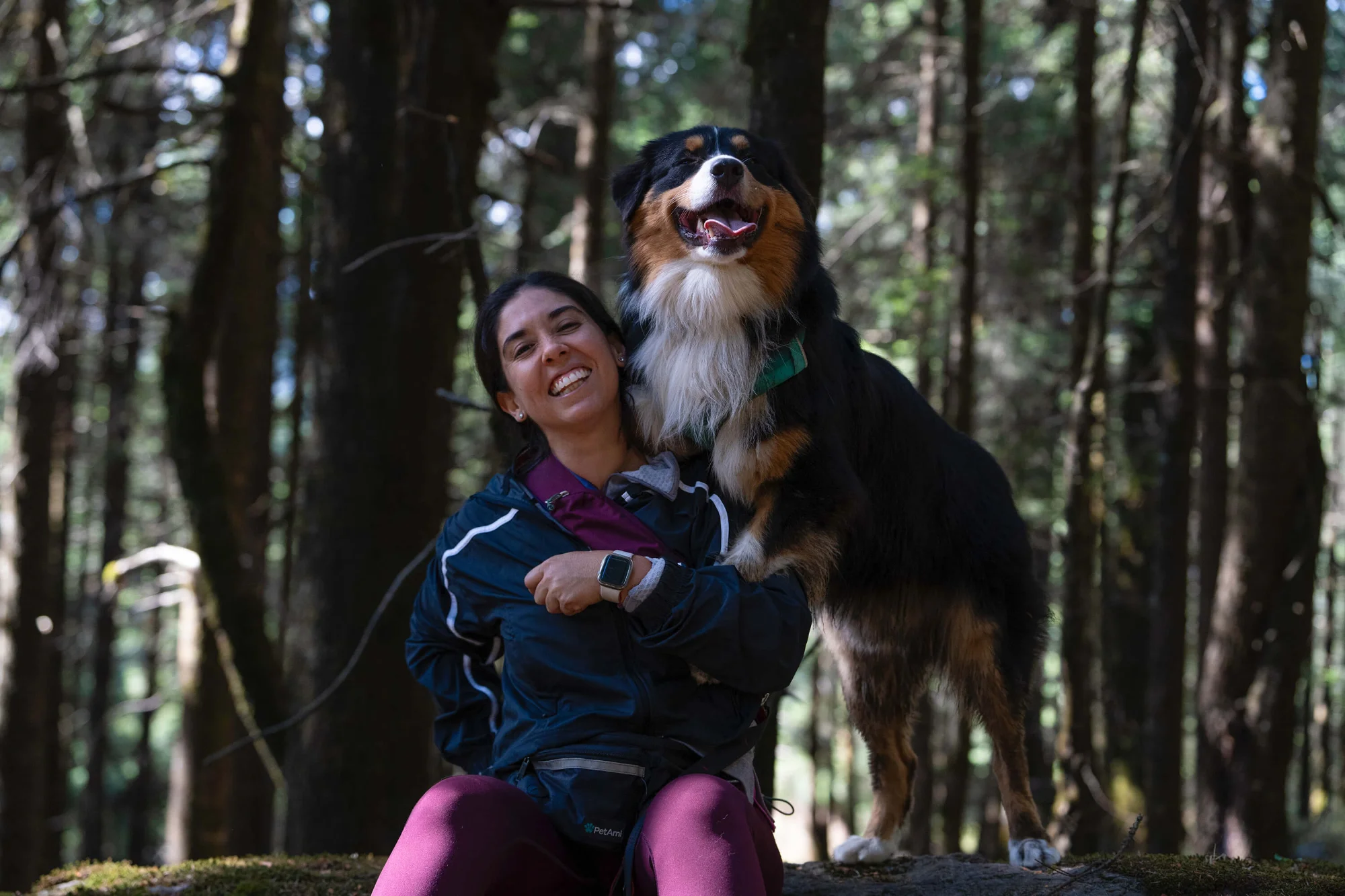 Una persona sonríe sentada en una roca en un bosque, con un perro grande y peludo a su lado. El perro apoya su pata en el hombro de la persona, y ambos parecen felices. La luz del sol se filtra entre los árboles.