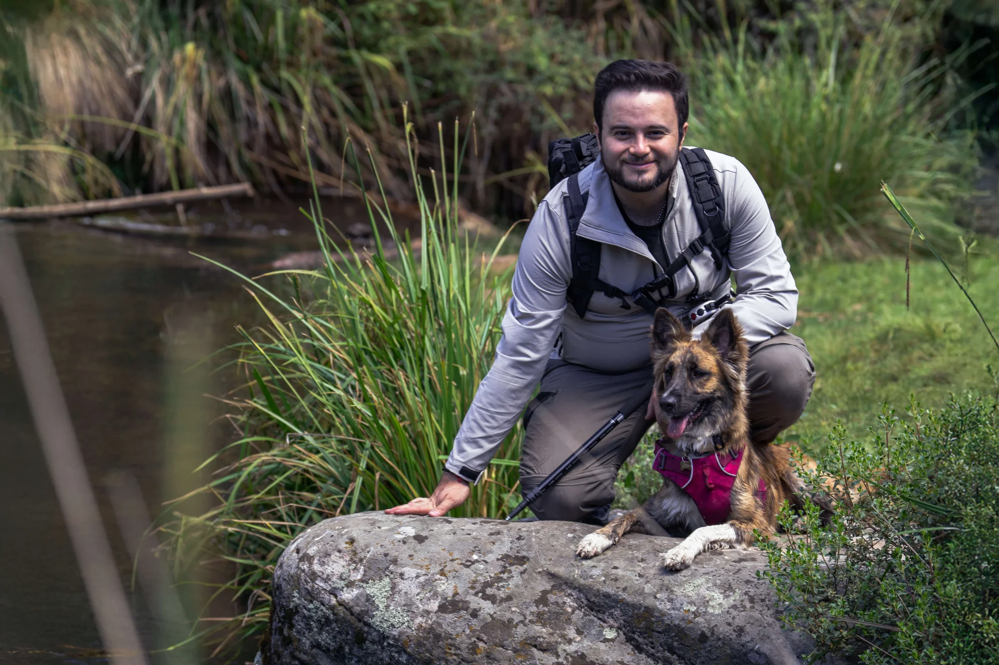 Un hombre con ropa de calle y una cámara en la mano se agacha junto a un río, apoyando la mano en una roca. Un perro con un arnés rosa está sentado a su lado. La hierba alta y un río tranquilo crean un entorno sereno y natural.